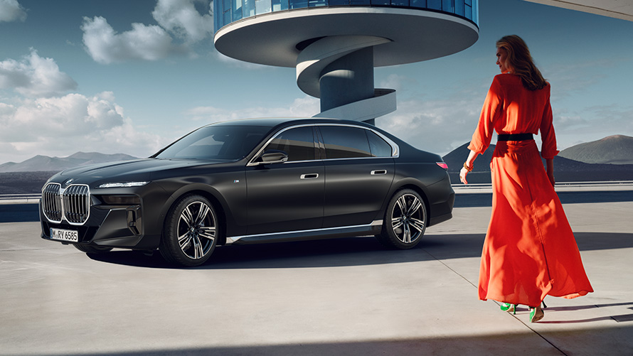 Model in red dress in front of BMW 7 series sedan G70 ICE M sports package BMW 7 Series Sedan G70 ICE M sports package Frozen Deep grey metallic 21" M light alloy wheels star spoke 908 three quarterFront view with model in red dress