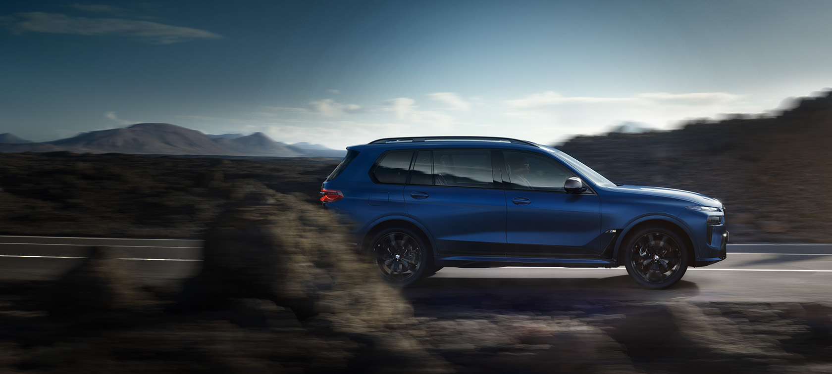 BWM X7 M60i xDrive G07 LCI Frozen Marina Bay Blue side view dirving on the street in front of mountain landscape